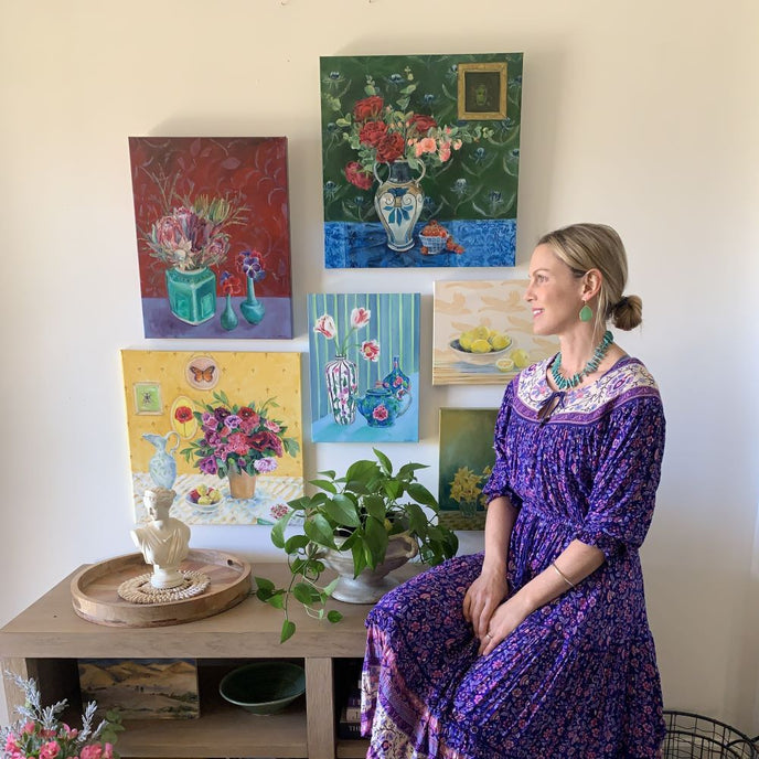  Artist Alana Clumeck posing in a purple dress next to a wall display of her vibrant floral paintings 
