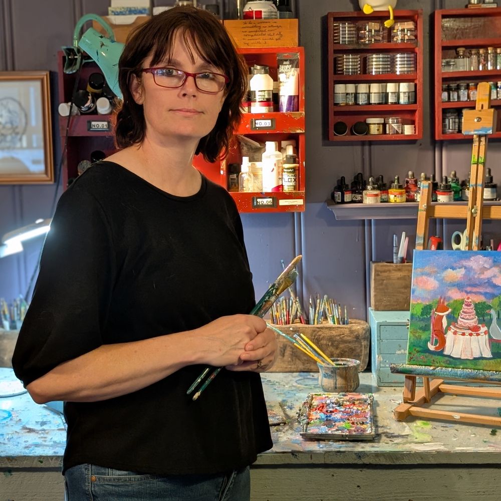  UGallery artist Andrea Doss standing in her studio holding paintbrushes beside a small painting on an easel, surrounded by shelves of art supplies 