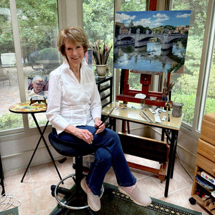  UGallery artist Faye Vander Veer sitting on a stool in a bright studio, holding a paintbrush beside an easel 
