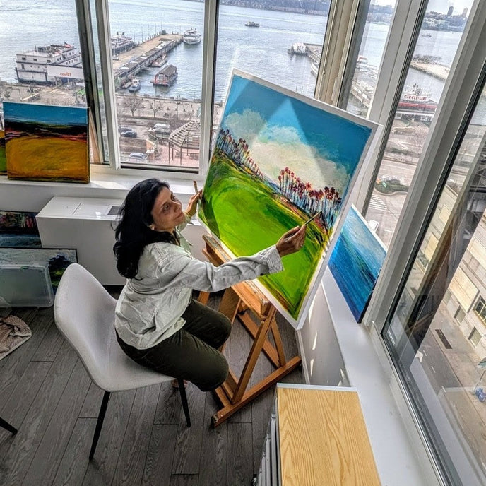  UGallery artist Kajal Zaveri painting a landscape on an easel beside large windows overlooking the Hudson River 