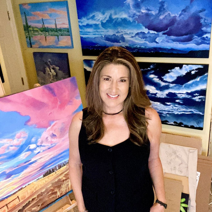  UGallery artist Renee Noel stands smiling in her studio filled with cloudscape paintings 